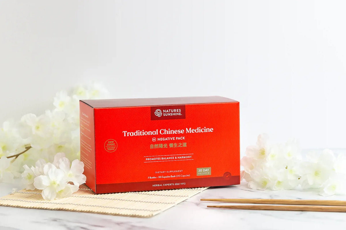 A red rectangular box of Nature's Pharmacy Traditional Chinese Medicine capsules, styled on a marble surface with white flowers and chopsticks.
