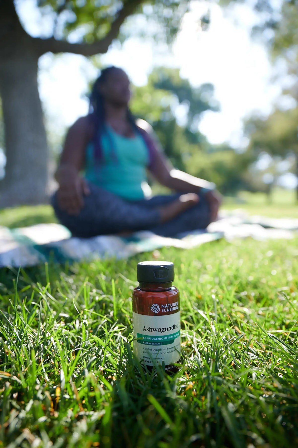 A small bottle of Nature's Sunshine Ashwagandha placed in green grass with a blurred person meditating outdoors in the background.