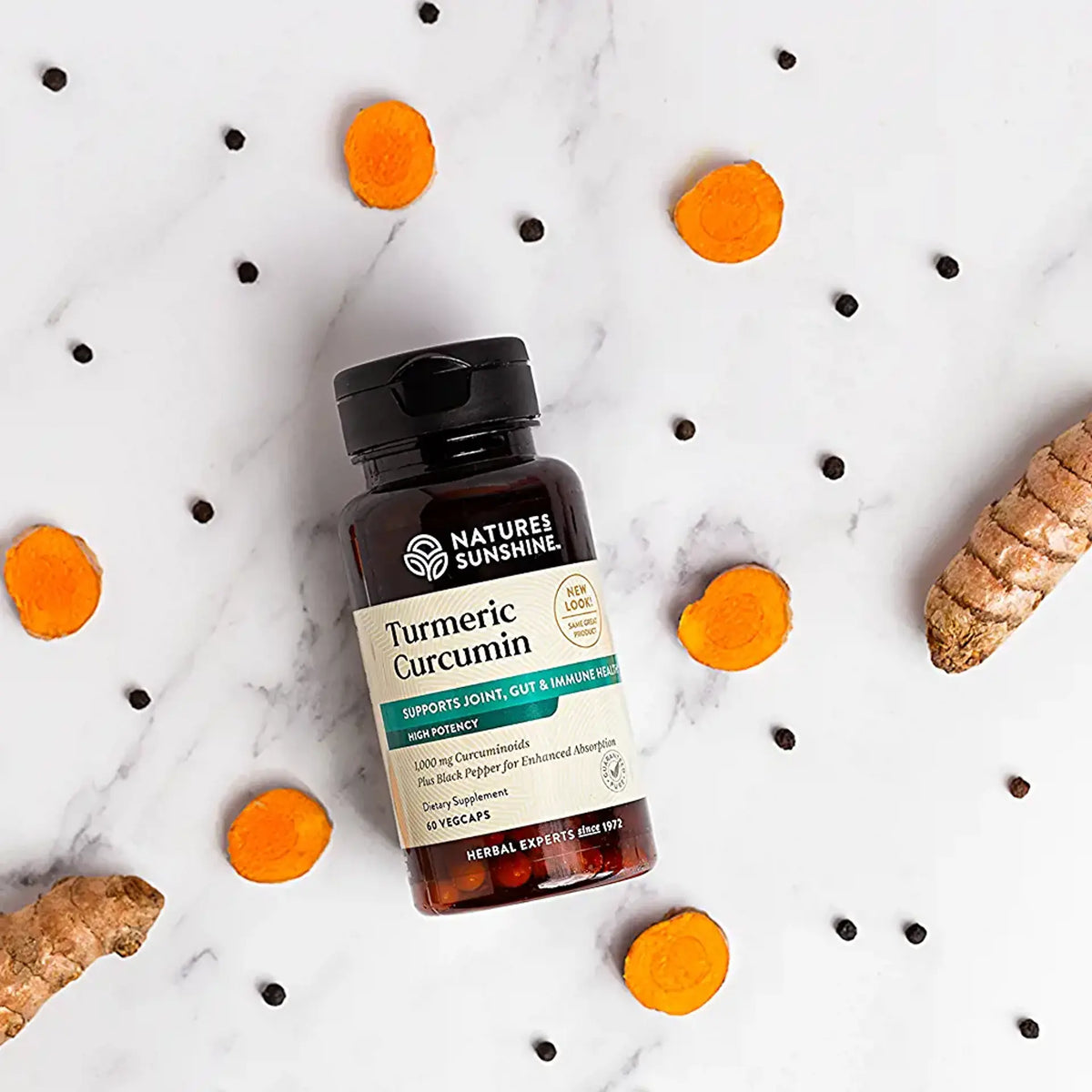 A bottle of Nature's Sunshine Turmeric Curcumin capsules arranged on a white marble background with scattered black peppercorns, sliced turmeric roots, and whole turmeric roots as styled props.