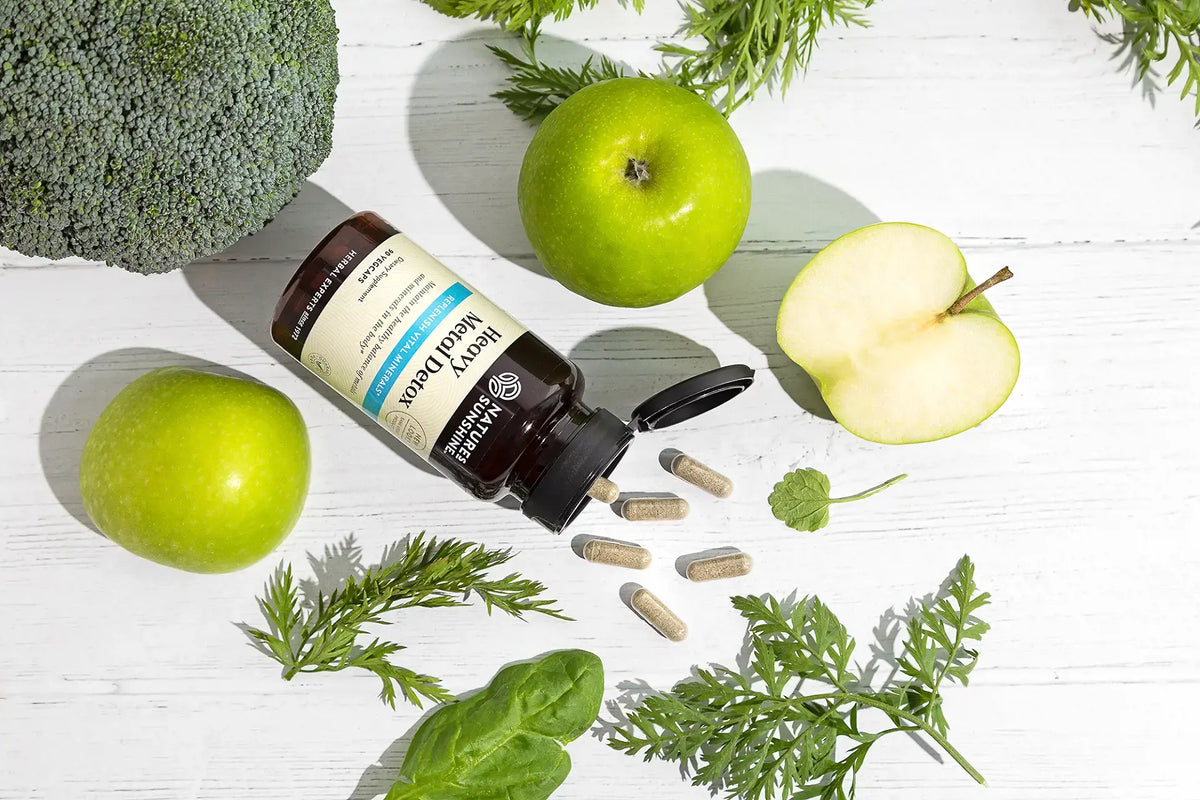 A bottle of Daily Health Foods Omega 3+ 2K supplement lies on its side on a white wooden surface, surrounded by green apples, a halved apple, broccoli, and leafy greens. Several capsules are scattered from the open bottle.