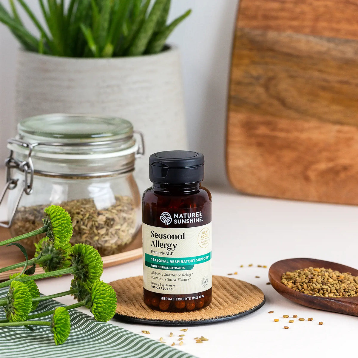 A bottle of Nature's Sunshine Seasonal Allergy Syrup placed on a woven coaster on a white table, surrounded by herbal ingredients, a potted plant, and a wooden cutting board.
