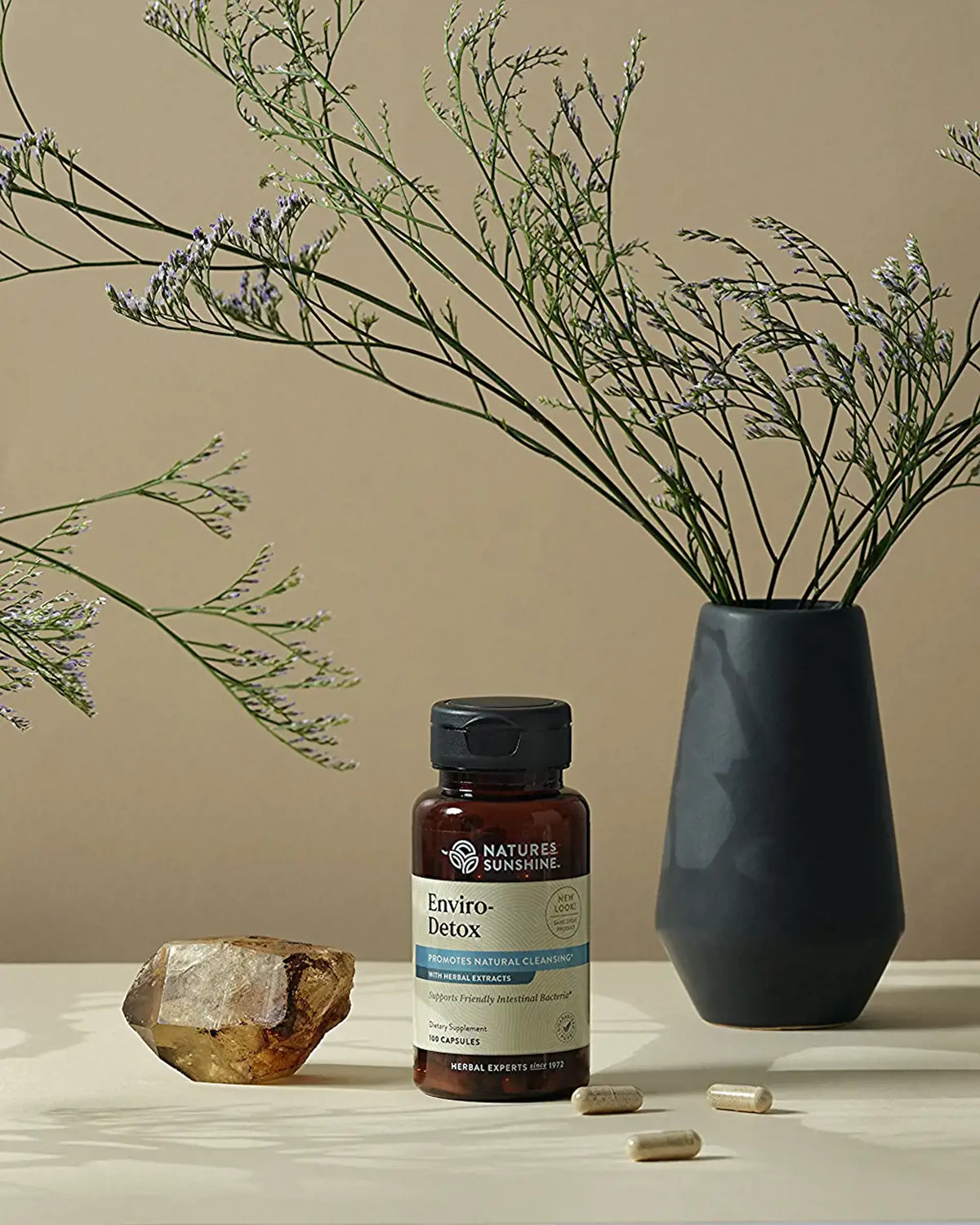 A bottle of Nature's Recommendations Enviro-Detox dietary supplement capsules, styled on a beige surface with a dark ceramic vase of dried stems, a crystal, and loose capsules.