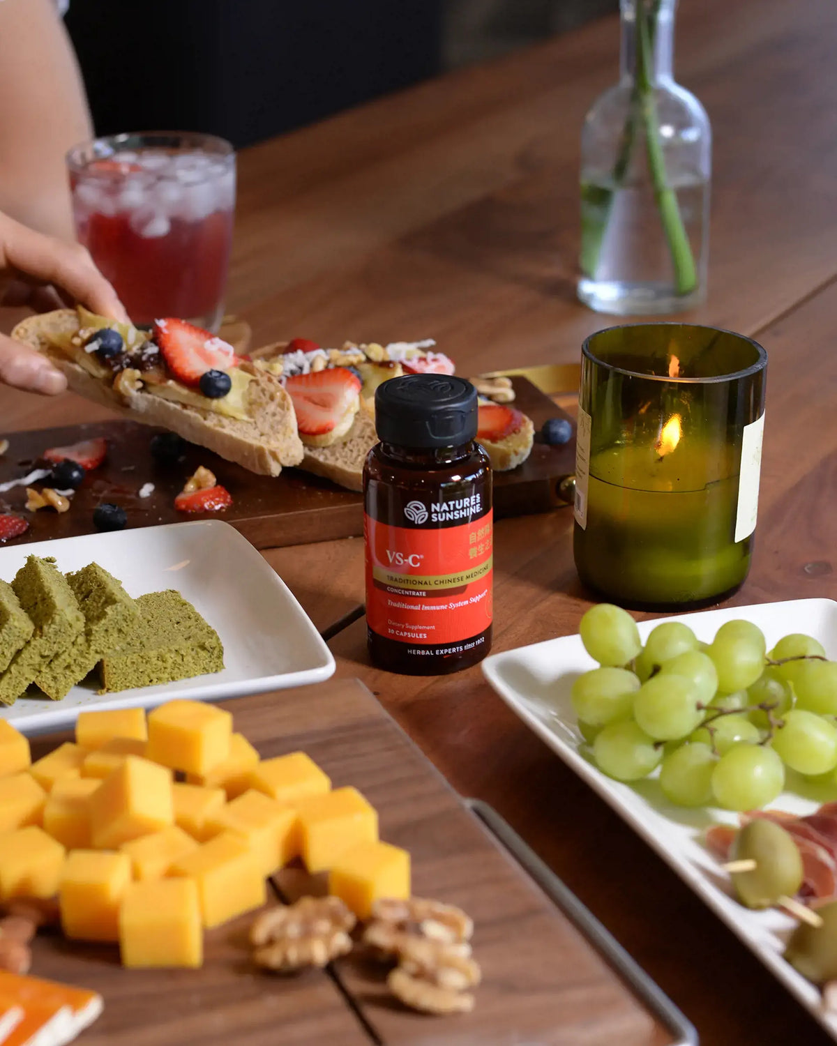 A bottle of Nature's Way Vitamin B3 is displayed on a wooden table surrounded by food, including green grapes, cheese cubes, crackers, and a hand holding a toasted sandwich in the background.