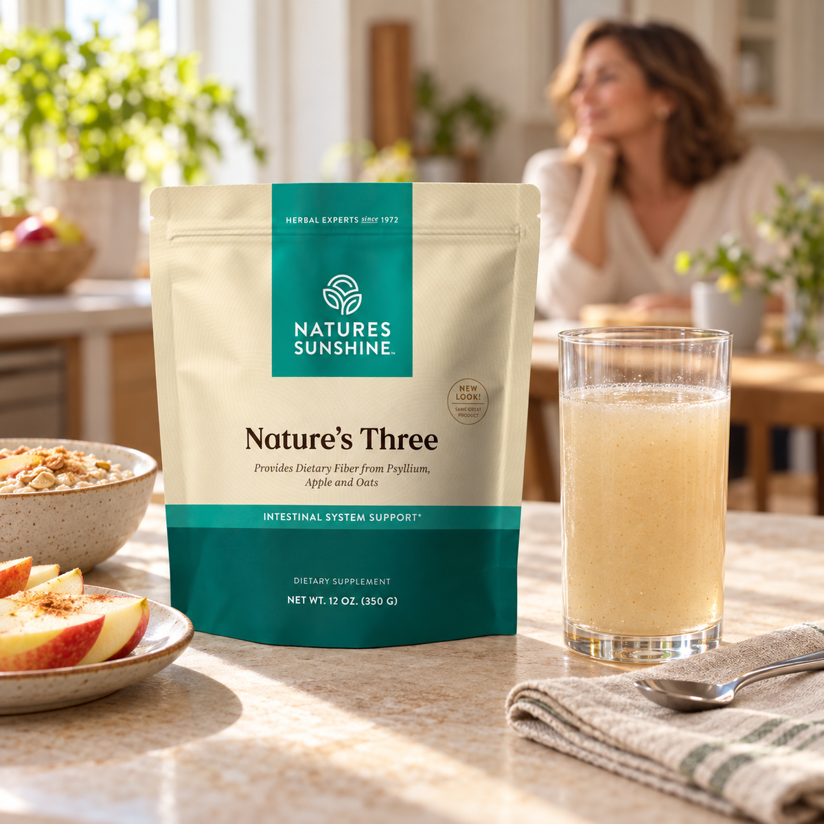Nature's Sunshine Nature's Three Intestinal System Support powder in a 12 oz stand-up pouch, displayed on a kitchen counter next to a glass of the prepared golden drink, with a woman in a warm, sunlit kitchen in the background.