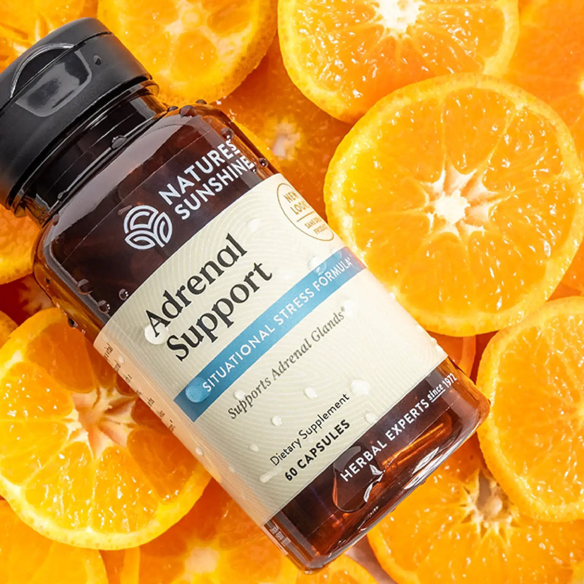 A bottle of Nature's Sunshine Adrenal Support dietary supplement placed diagonally on a background of fresh orange slices. The bottle is dark amber glass with a black cap and a cream-colored label featuring blue and gray text.