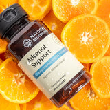 A bottle of Nature's Sunshine Adrenal Support dietary supplement placed diagonally on a background of fresh orange slices. The bottle is dark amber glass with a black cap and a cream-colored label featuring blue and gray text.