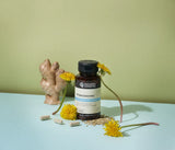 A bottle of Nature's Sunshine Prooctazyme digestive supplement, styled with yellow dandelions, a piece of fresh ginger root, and capsules on a light blue surface against a sage green background.