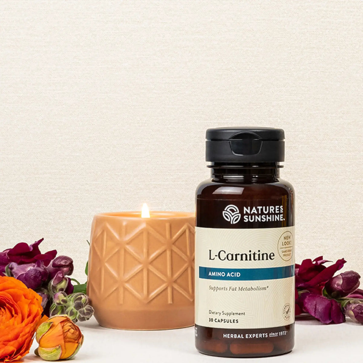 A bottle of Nature's Sunshine L-Carnitine dietary supplement stands on a white surface next to a lit beige candle, with orange and purple flowers arranged around it against a textured off-white background.