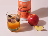 A glass of iced orange-toned herbal tonic with ice cubes and dark fruit pieces, placed beside a red apple and a sliced apple on a pale pink surface. A white bottle labeled Natures Sunshine Chinese Mineral-Chi Tonic stands behind the drink.