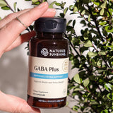 Nature's Sunshine GABA Plus supplement bottle held by a hand with a blurred green plant background