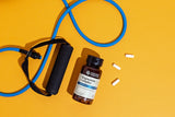 A bottle of Magnesium Complex supplement by Nature's Answers lies on a bright yellow background next to a blue resistance band and three white capsules.