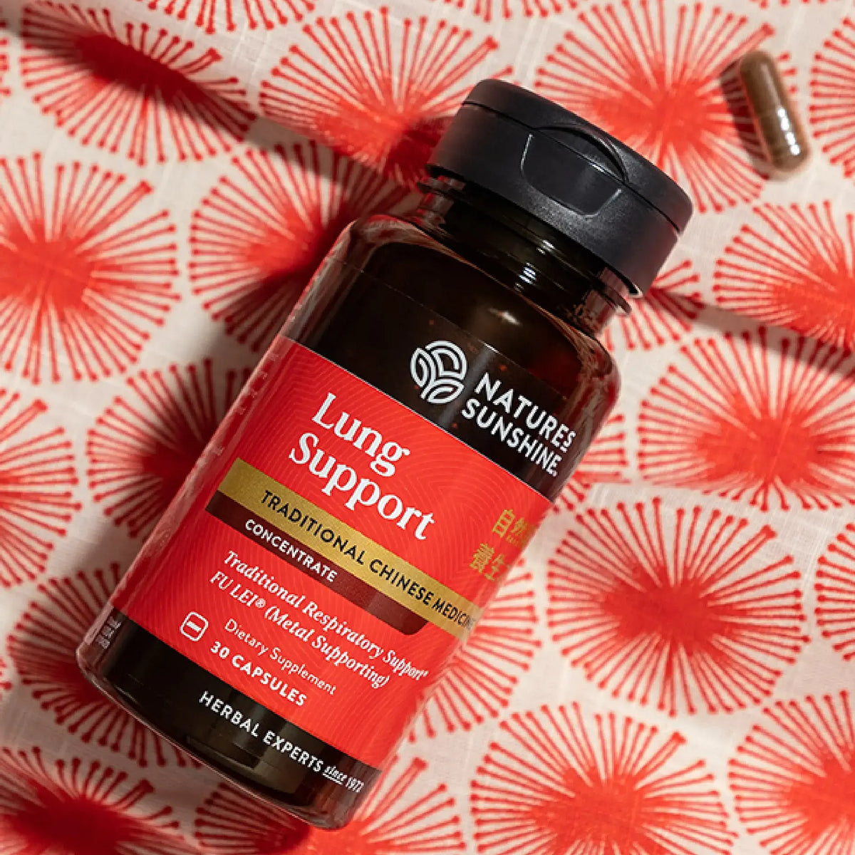 A bottle of Nature's Sunshine Lung Support dietary supplement capsules, placed on a red and white patterned fabric background with a single capsule nearby.