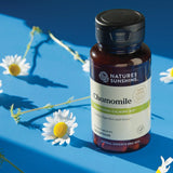 A bottle of Nature's Sunshine Chamomile capsules placed on a bright blue surface with scattered chamomile flowers around it.