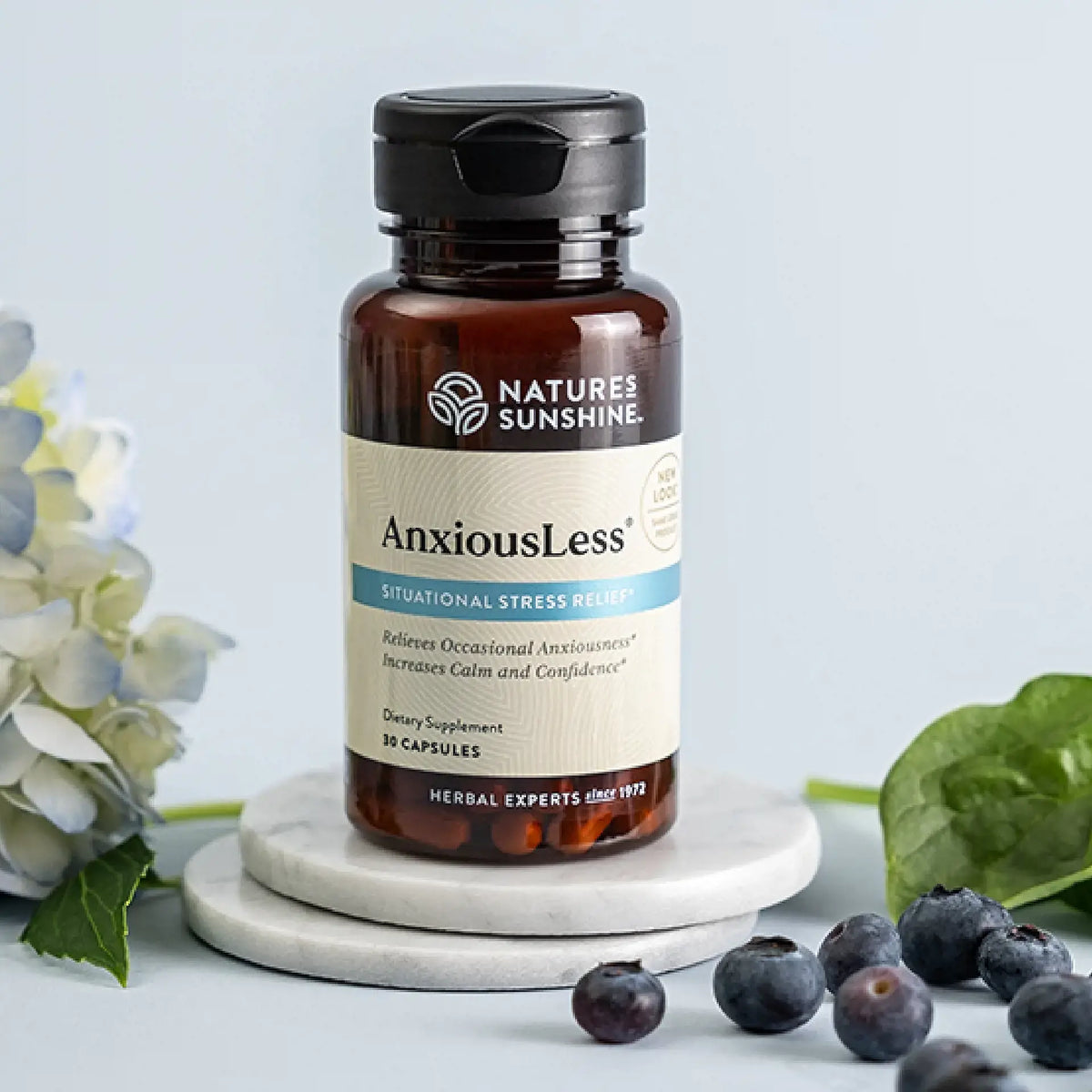 Nature's Sunshine AnxiousLess dietary supplement bottle on a white marble coaster, styled with blueberries, spinach leaves, and a white flower on a light blue background.