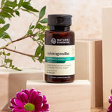 A bottle of Nature's Sunshine Ashwagandha herbal extract supplement, displayed on a light wooden block with decorative green leaves and pink flowers in the background.