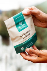 A close-up of a hand holding a stand-up pouch of Nature's Sunshine MyImmune Defense dietary supplement. The pouch is cream-colored with teal-green bands at the top and bottom, and black text.