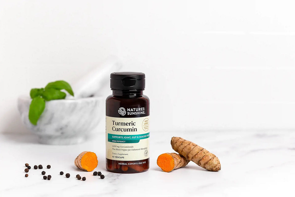 A bottle of Nature's Sunshine Turmeric Curcumin capsules on a white marble surface, styled with fresh turmeric roots, sliced turmeric showing its bright orange interior, scattered black peppercorns, and a blurred marble mortar and pestle with green herbs in the background.