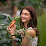 A smiling woman with long brown hair stands outdoors among green leaves, holding a small orange tube package in both hands.