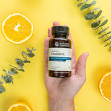 Hand holding a bottle of Nature's Sunshine Vitamin C supplement on a yellow background with oranges and eucalyptus leaves.