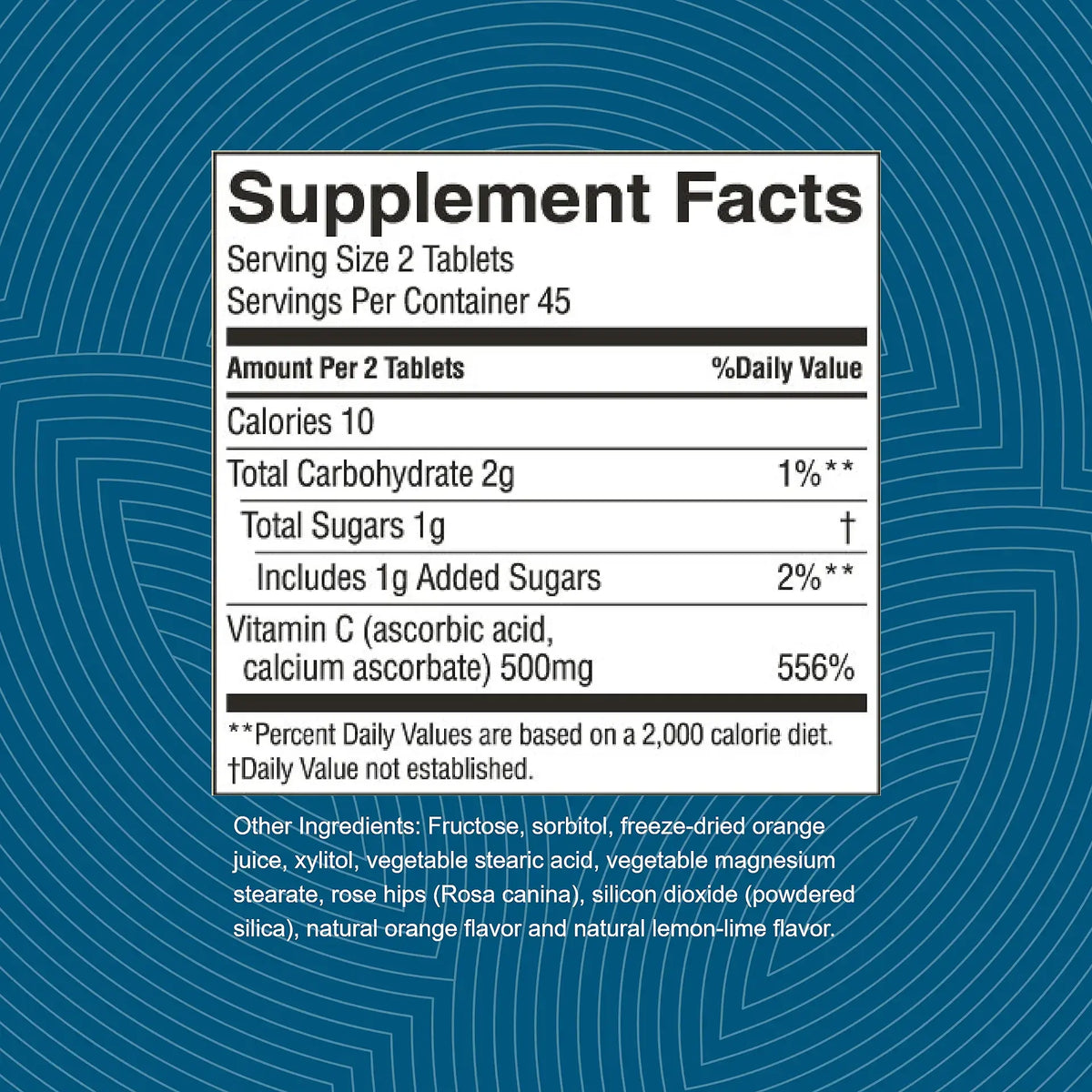 A supplement facts label shown on a blue patterned background. The label reads 'Supplement Facts' with serving size 2 tablets, calories 10, total carbohydrate 2g, total sugars 1g, includes 1g added sugars, vitamin C 556%, and lists other ingredients including fructose, sorbitol, vegetable stearic acid, vegetable magnesium stearate, rose hips, silicon dioxide, natural orange flavor, and natural lemon-lime flavor.