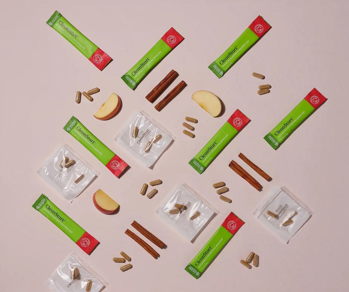 Flat-lay of several green and red Herbalinfusion stick packs arranged on a pale pink background, with cinnamon sticks, apple slices, and small clear packets containing capsules around them.