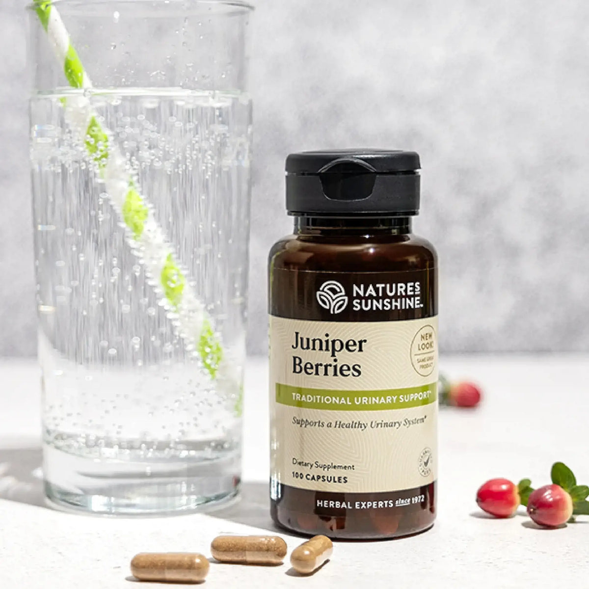 A bottle of Nature's Sunshine Juniper Berries dietary supplement stands on a light surface next to a glass of water with a green-and-white striped straw, with a few capsules and red berries around it.