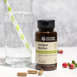 A bottle of Nature's Sunshine Juniper Berries dietary supplement stands on a light surface next to a glass of water with a green-and-white striped straw, with a few capsules and red berries around it.