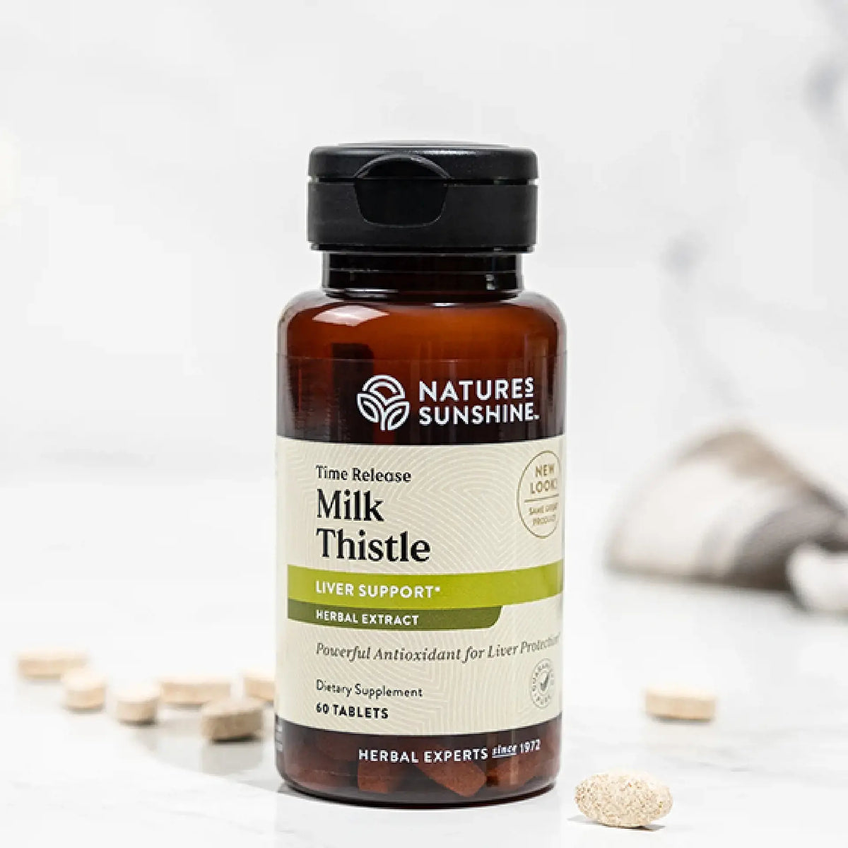 A bottle of Nature's Sunshine Milk Thistle herbal supplement with a black cap and amber-brown bottle, shown on a light marble surface with a few tablets scattered around it.
