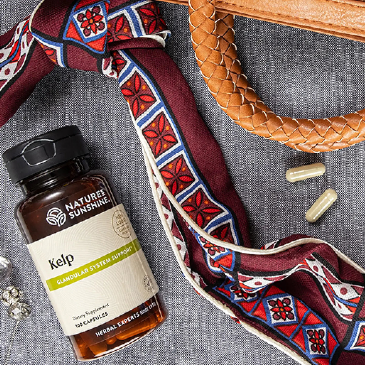 A bottle of Nature's Sunshine Kelp dietary supplement lies on a textured gray fabric surface, styled with a red, blue, and white patterned silk scarf, a brown woven strap, and two capsules.