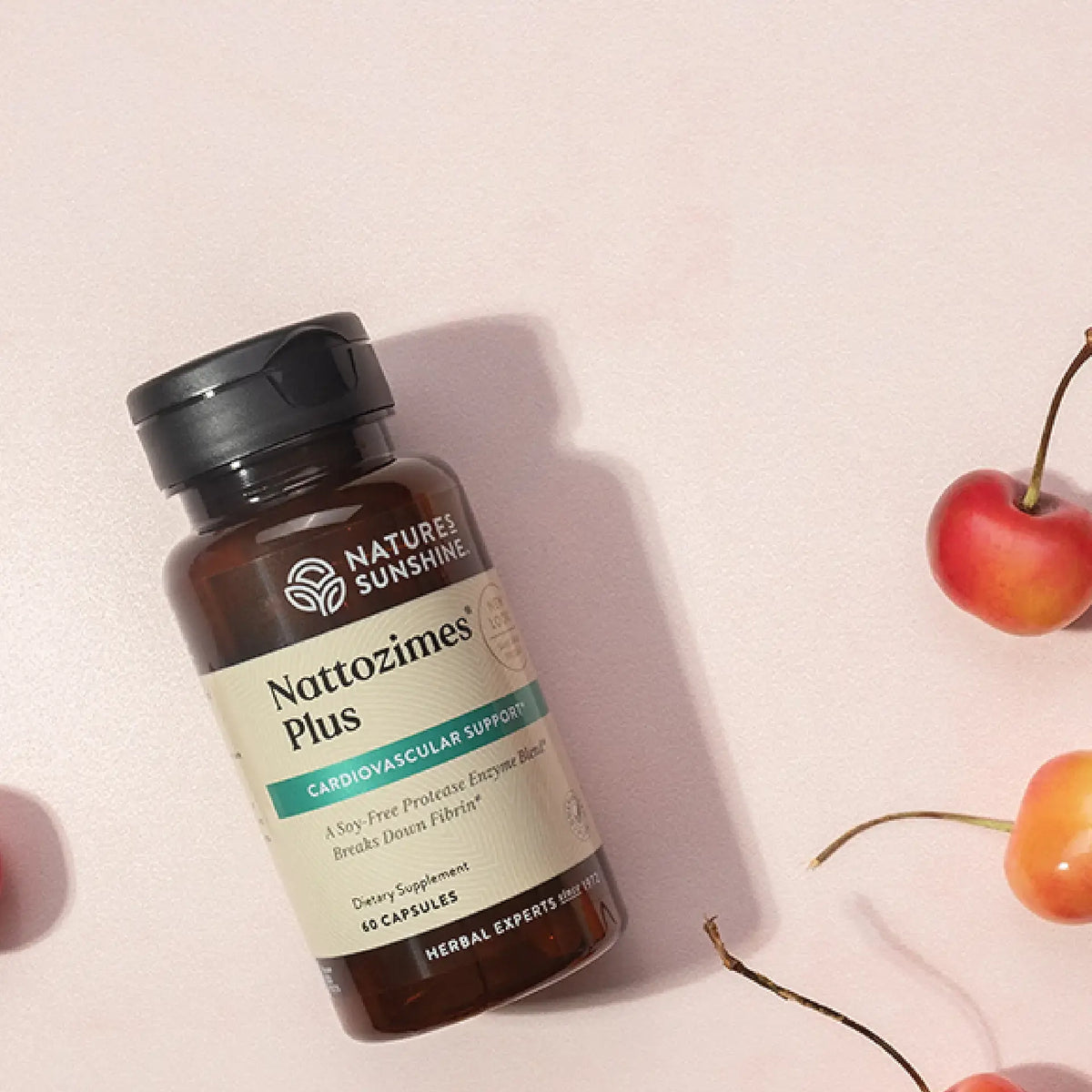 A bottle of Nature's Sunshine Nattozimes Plus dietary supplement capsules, styled on a light pink background with decorative cherries.