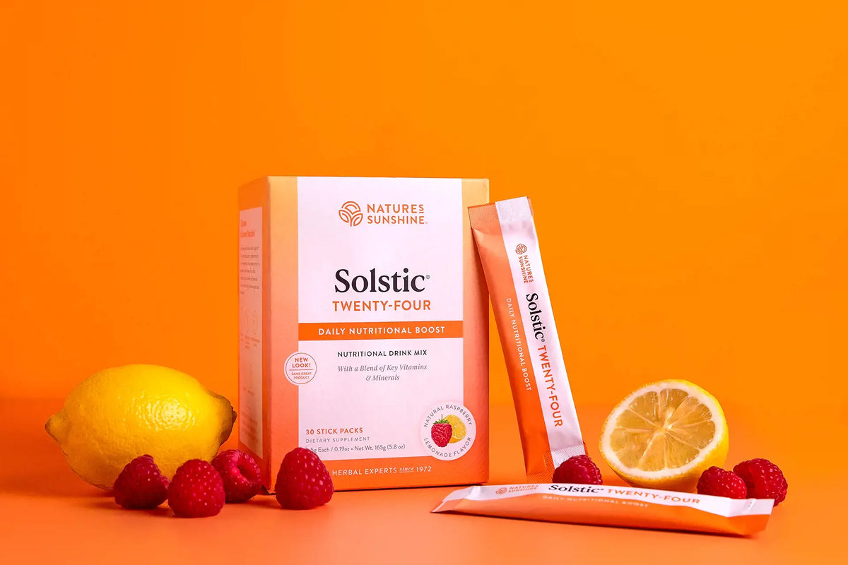 Nature's Sunshine Solstic Twenty-Four daily nutritional drink mix box with two single-serve stick packs, styled with fresh raspberries, a whole lemon, and a sliced lemon on a bright orange background.