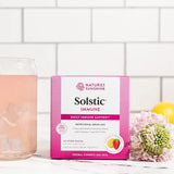 A box of Nature's Sunshine Solstic Immune herbal drink mix stands on a white surface against a white subway tile wall. A pink beverage sits in a glass on the left, and flowers and a lemon are placed on the right.
