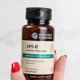 A hand with red painted nails holds a bottle of Nature's Sunshine APS-II dietary supplement against a light gray background.