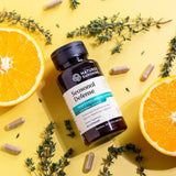 A bottle of Nature's Sunshine Seasonal Defense herbal supplement capsules, styled in a flat-lay on a bright yellow background with orange slices, fresh thyme, and scattered capsules.