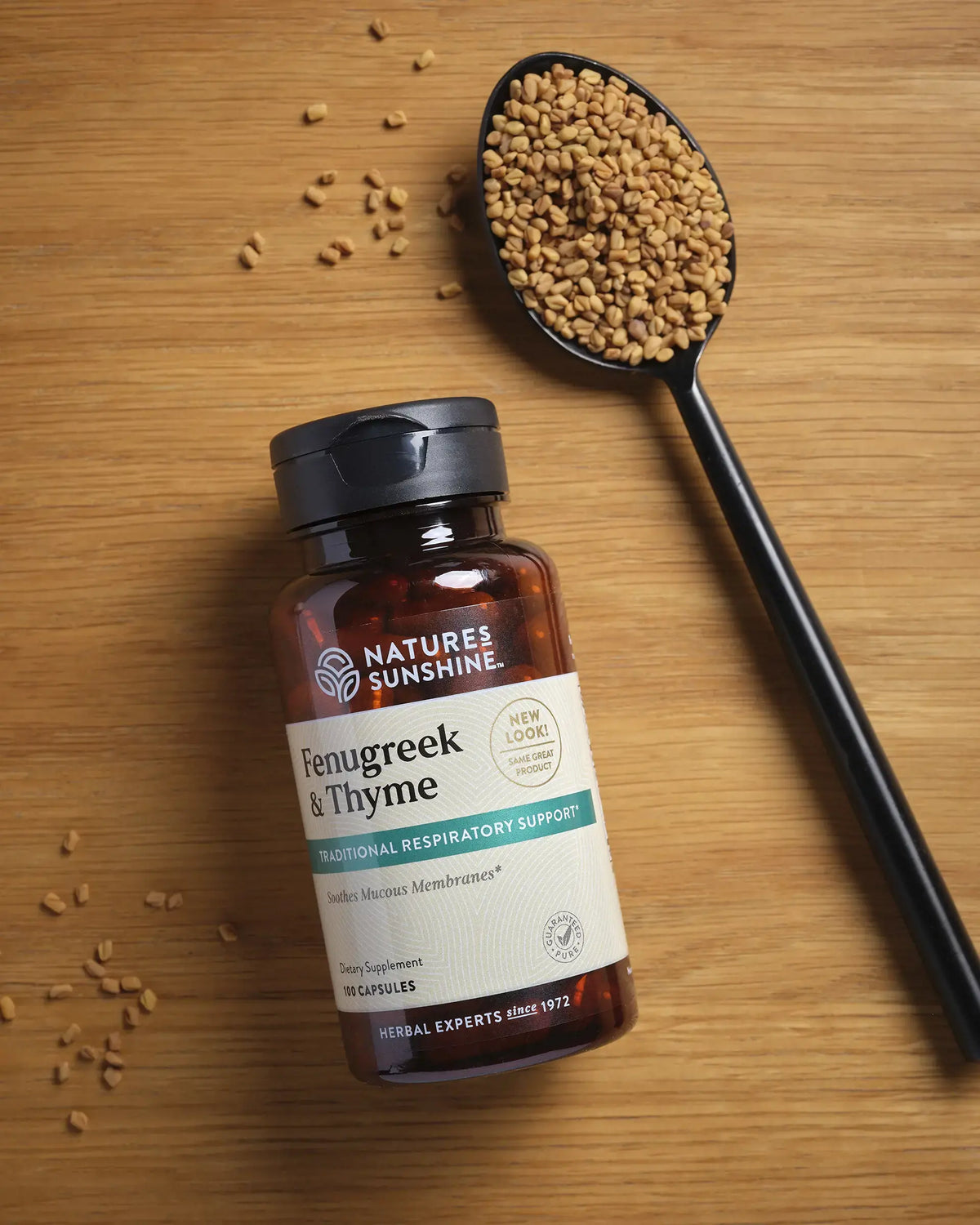 A bottle of Nature's Sunshine Fenugreek & Thyme capsules placed on a wooden surface next to a black spoon filled with fenugreek seeds.