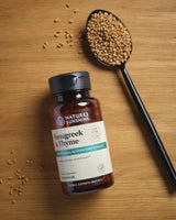 A bottle of Nature's Sunshine Fenugreek & Thyme capsules placed on a wooden surface next to a black spoon filled with fenugreek seeds.