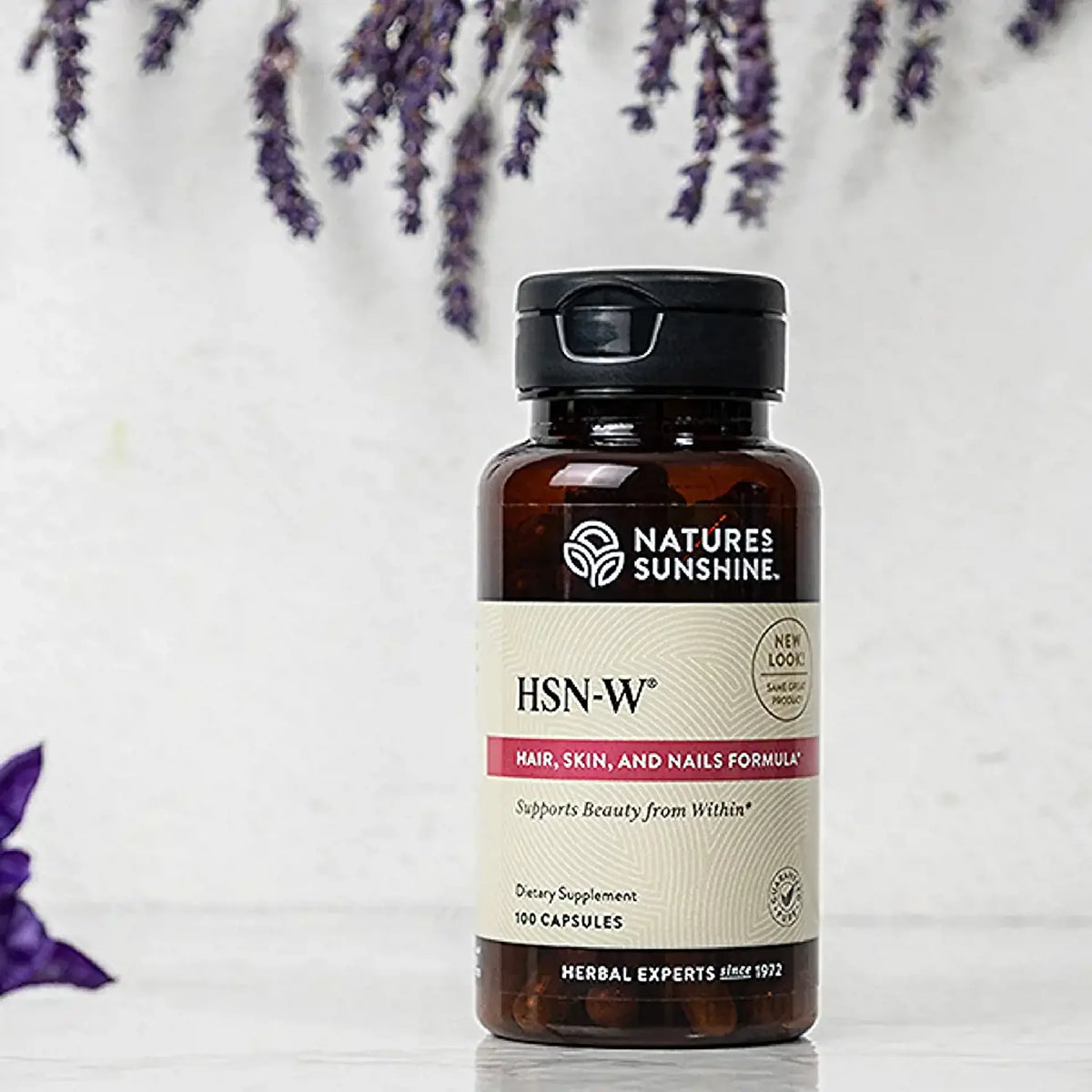 A bottle of Natures Sunshine HSN-W Hair, Skin, and Nails Formula dietary supplement, featuring an amber glass bottle with a black cap and cream-colored label, set against a white background with decorative purple flowers.