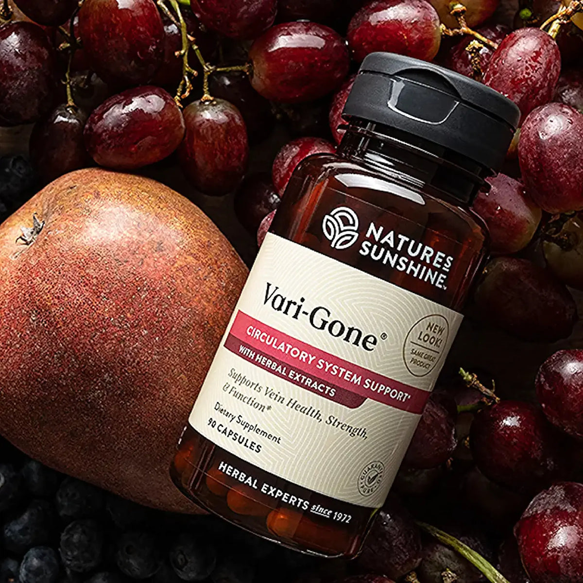 A bottle of Natures Sunshine Vari-Gone dietary supplement, surrounded by fresh red grapes and a pomegranate. The amber glass bottle has a black cap and a white label with red and black text.