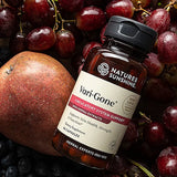 A bottle of Natures Sunshine Vari-Gone dietary supplement, surrounded by fresh red grapes and a pomegranate. The amber glass bottle has a black cap and a white label with red and black text.