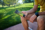 A man sitting outdoors in a park holding a tube of NATURE'S SUNSHINE  Tei-fu Lotion product, wearing a yellow t-shirt and a black smartwatch.