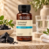 A bottle of Nature's Sunshine Activated Charcoal dietary supplement, 120 capsules, displayed on a stone surface with charcoal pieces, a glass of water, and a towel in the background.