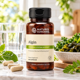 A bottle of Nature's Sunshine Algin dietary supplement stands on a marble countertop with a glass of water, loose capsules, and green herbs in a bright kitchen setting.