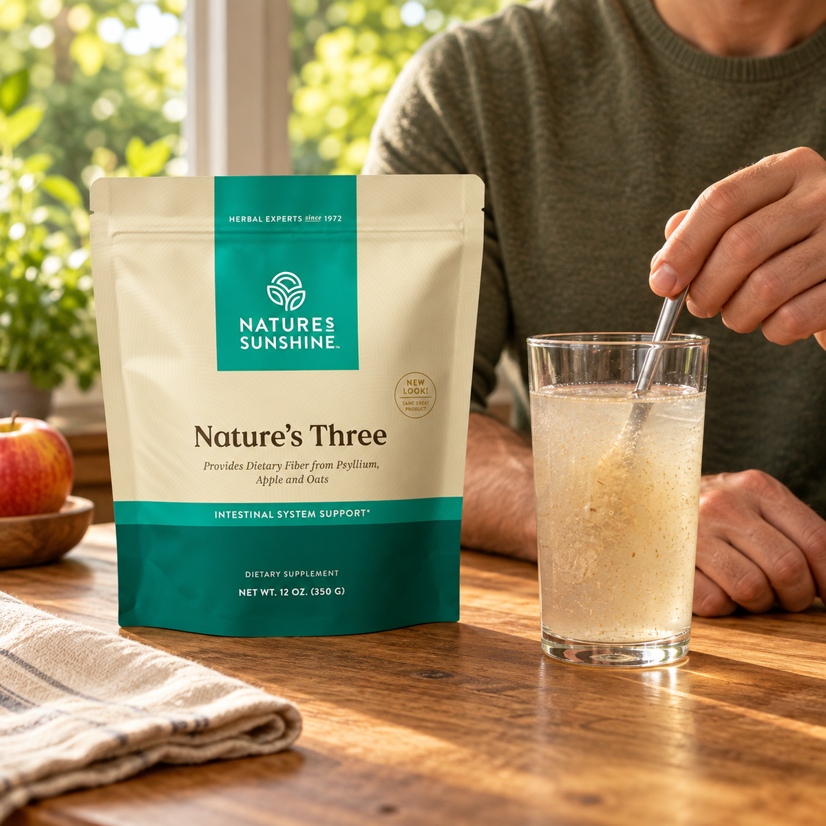 Nature's Sunshine Nature's Three dietary supplement pouch on a wooden table next to a glass of herbal tea being stirred by a person in a kitchen setting.