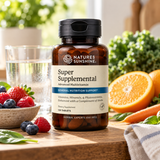 A bottle of Nature's Sunshine Super Supplemental dietary vitamins stands on a wooden table surrounded by fresh fruits and vegetables, including strawberries, blueberries, an orange, and leafy greens.