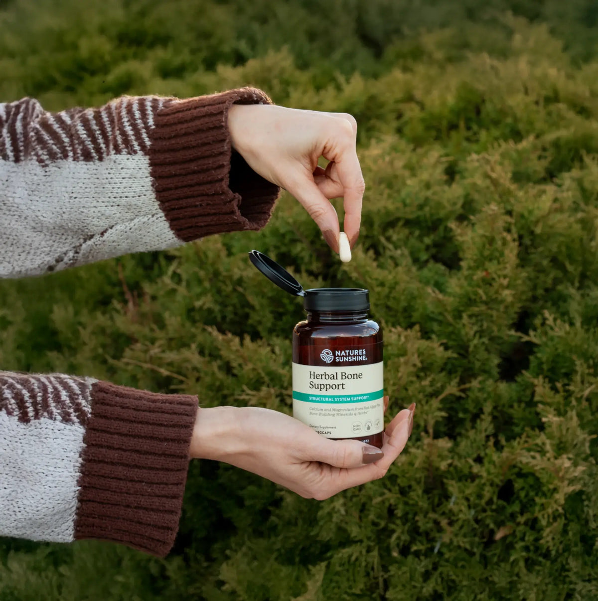 Person holding a bottle of Nature's Sunshine Herbal Bone Support supplement outdoors.