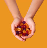 Two hands holding a small pile of colorful translucent gummy candies against a solid orange background.