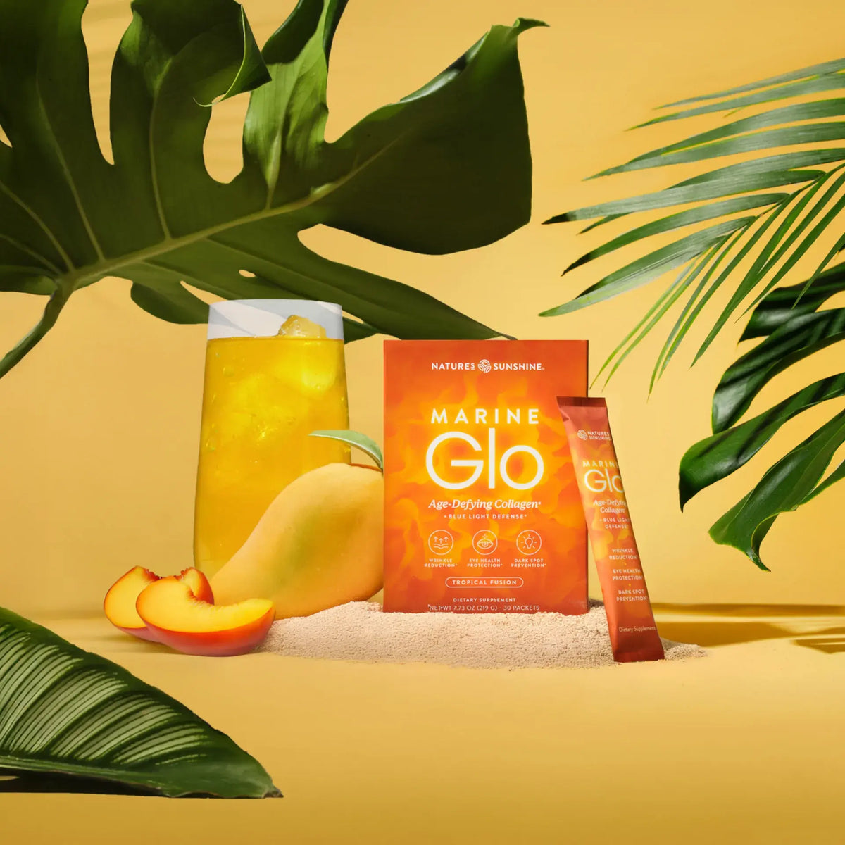 Nature's Sunshine Marine Glo dietary supplement box and sachet displayed with a glass of orange drink, sliced mango, and tropical leaves on a yellow background.